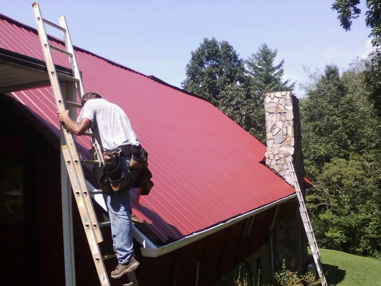 Roofer on a ladder beside a red metal roof and stone chimney — Soffit & Fascia Repair by Roofing Brothers in Old Westbury